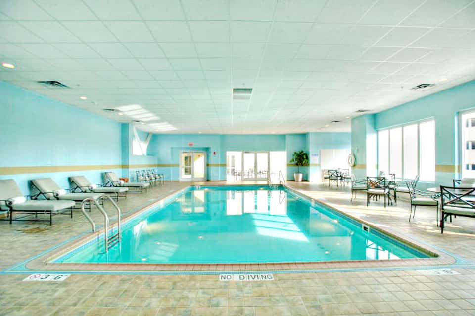 Indoor lap pool with turquoise water, lounge chairs, and large windows in a bright aquatic facility