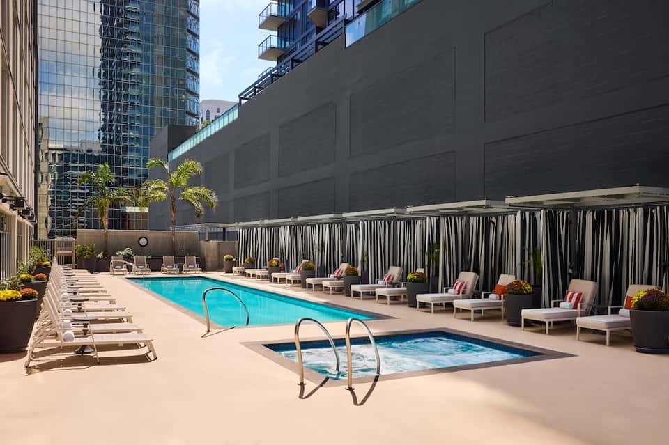 Urban hotel pool deck with lounge chairs, hot tub, and city building views with striped privacy screens