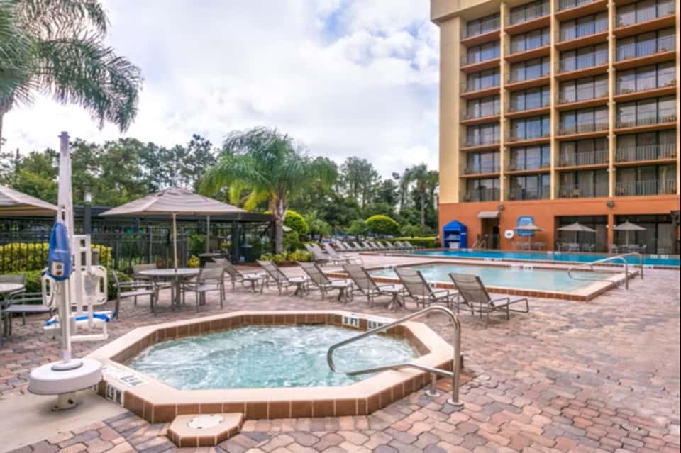 Resort pool and hot tub area with lounge chairs, gazebo, palm trees, and hotel building