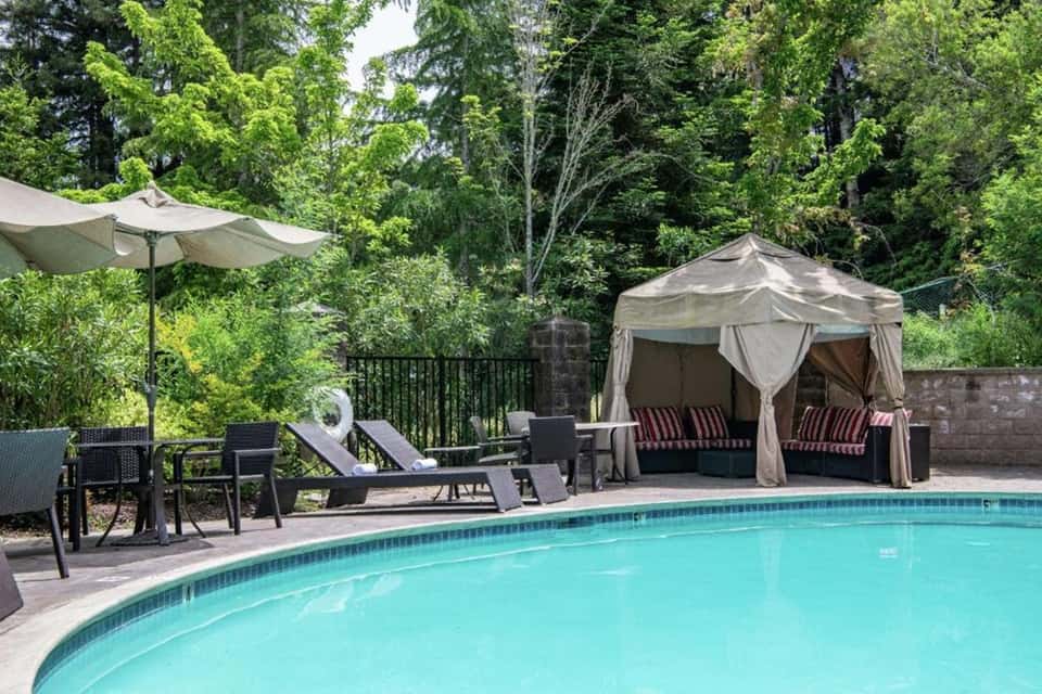 Resort pool area with cabana, lounge chairs, umbrellas, and wooded backdrop