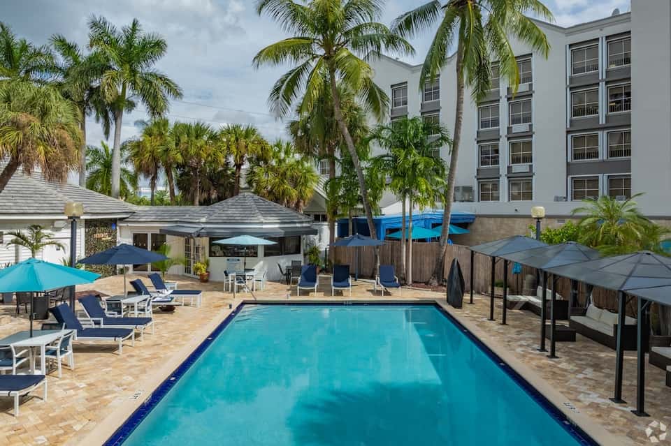 Resort pool with turquoise water, palm trees, lounge seating areas, cabanas, and multi-story hotel building in background