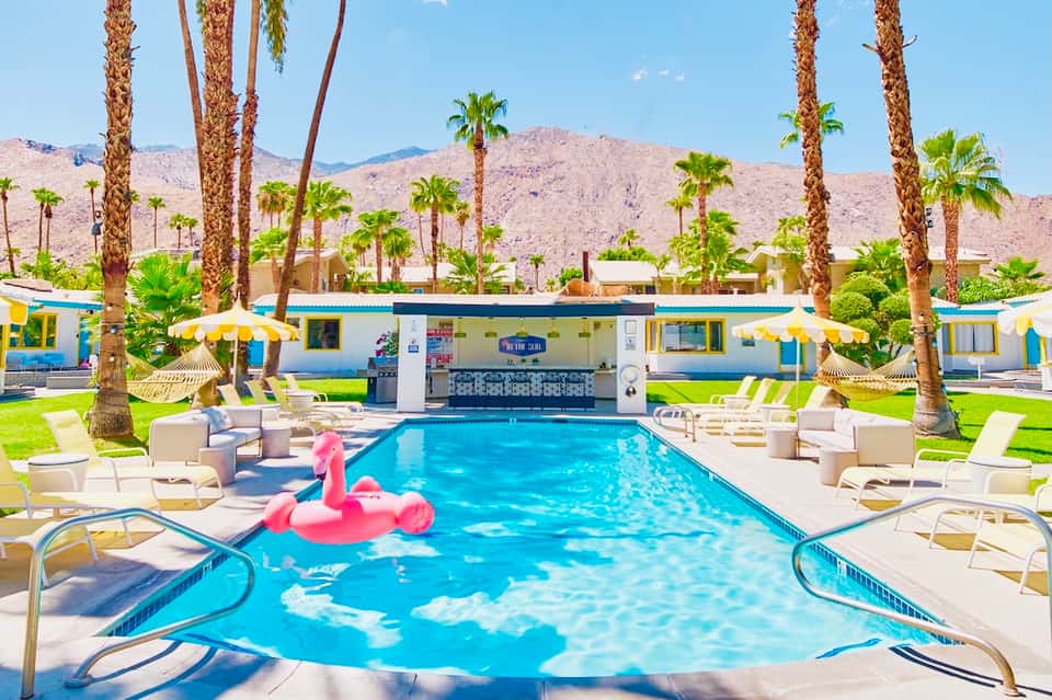 Resort pool with pink flamingo float, palm trees, lounge chairs, and desert mountain backdrop