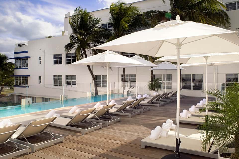 Resort pool deck with lounge chairs, large white umbrellas, palm trees, and modern hotel building