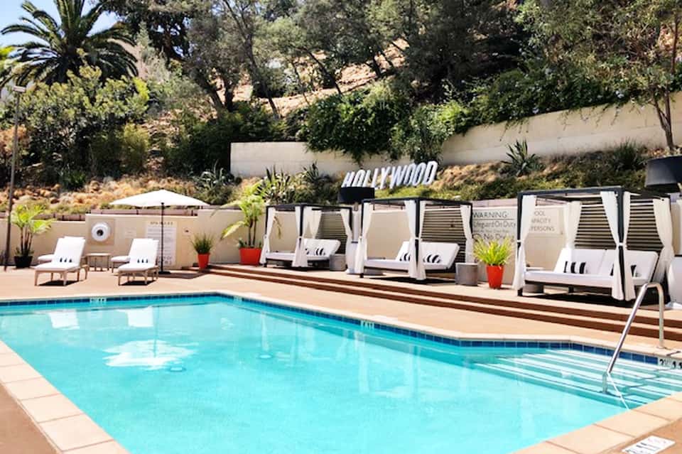 Resort swimming pool with white cabanas, lounge chairs, and hillside views under clear sky