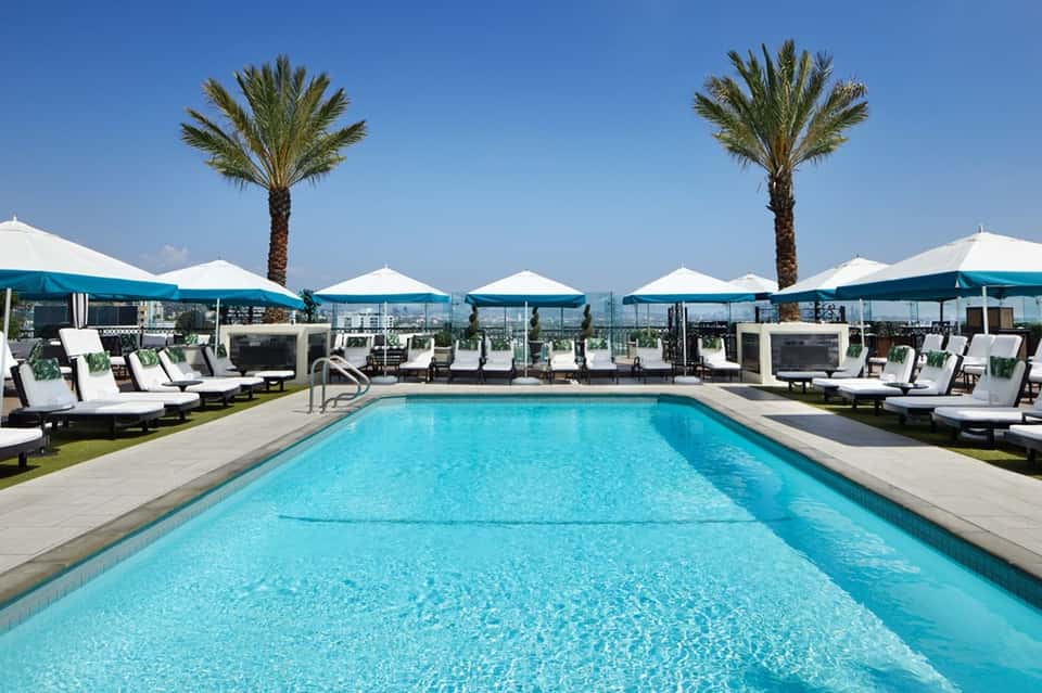 Resort pool with blue umbrellas, lounge chairs, palm trees, and mountain city skyline view