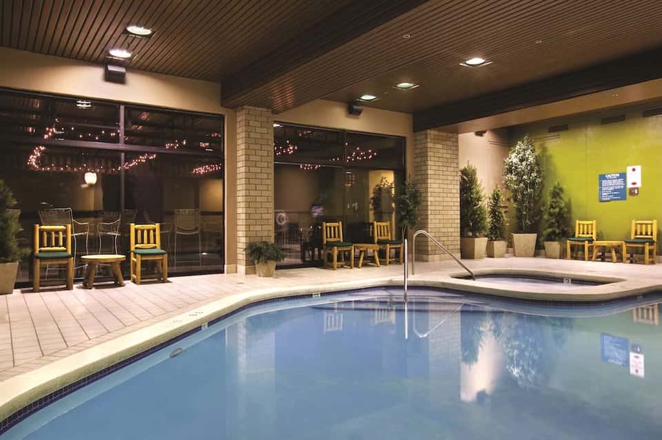 Indoor pool facility with turquoise water, tile decking, yellow seating, warm lighting, and glass doors to outdoor space
