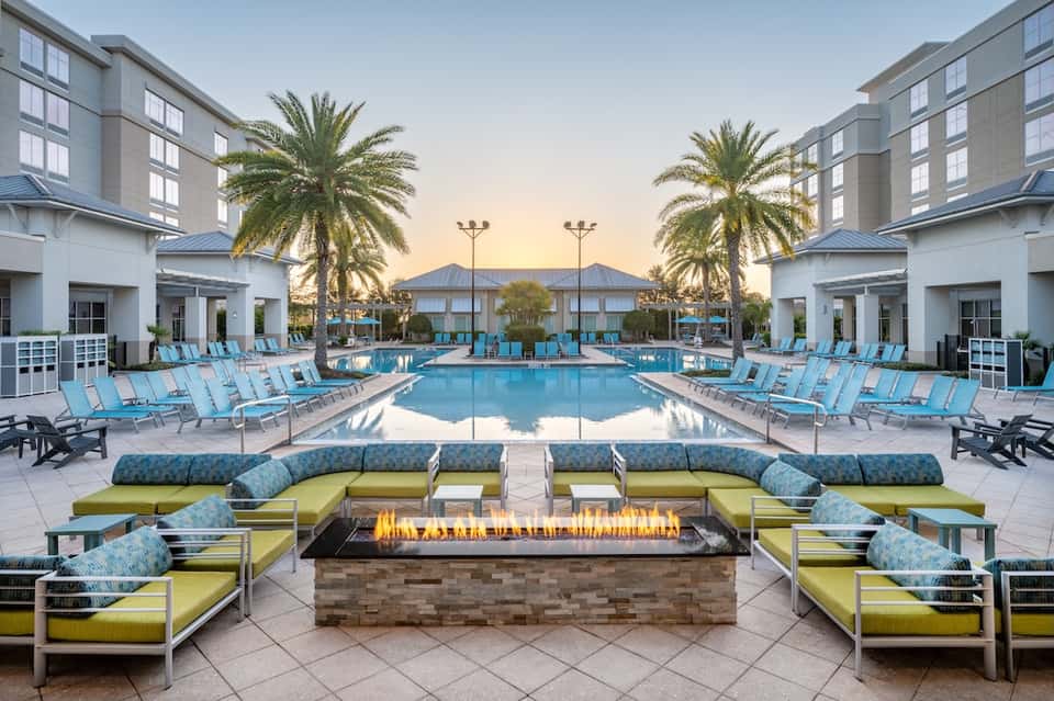 Resort pool deck at dusk with turquoise loungers, lime cushioned seating, fire pit, palm trees, and modern buildings