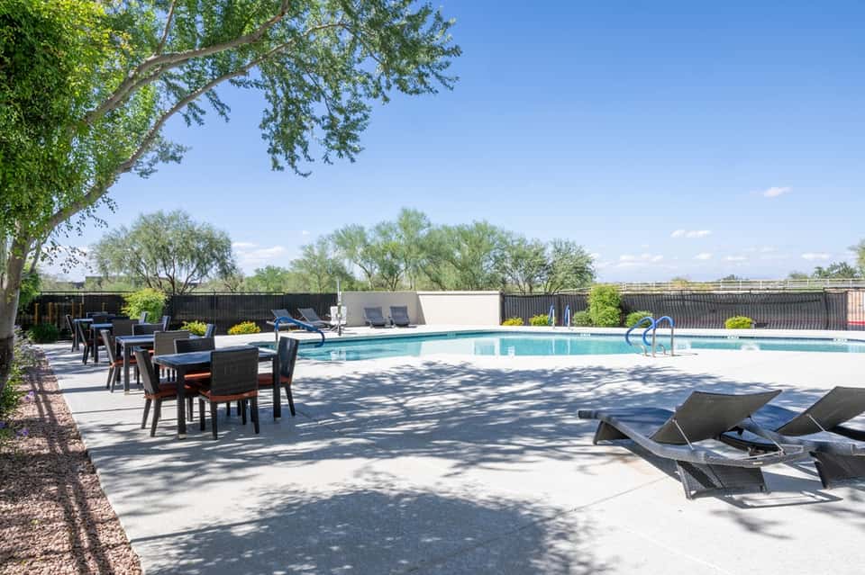 Resort pool area with shaded seating, lounge chairs, mature trees, and fenced grounds