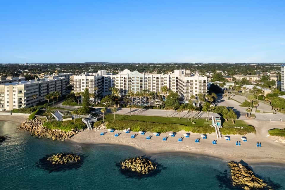 Beachfront resort with multi-story buildings, sand, blue umbrellas, and ocean view