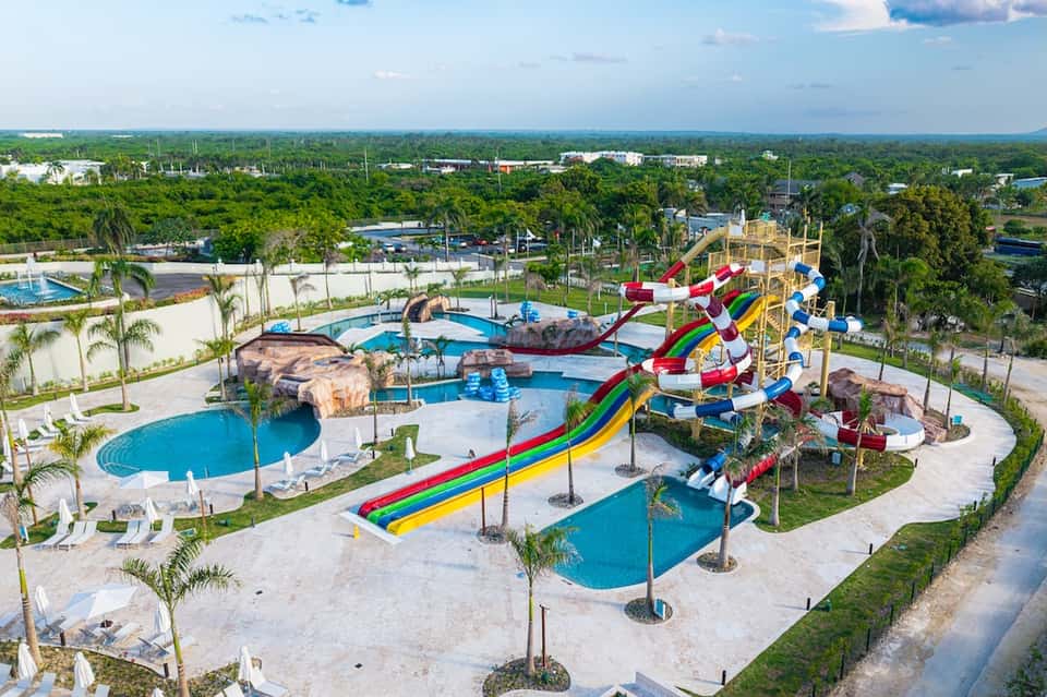 Aerial view of water park with colorful slides, multiple pools, palm trees, and surrounding forested landscape