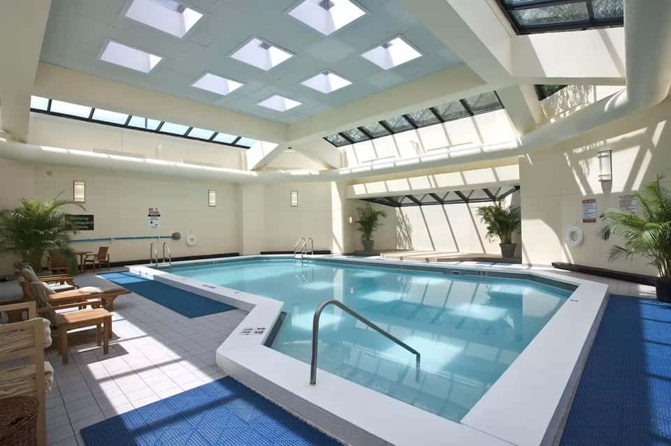 Bright indoor pool facility with skylights, spa tub, lounge seating, and tropical plants