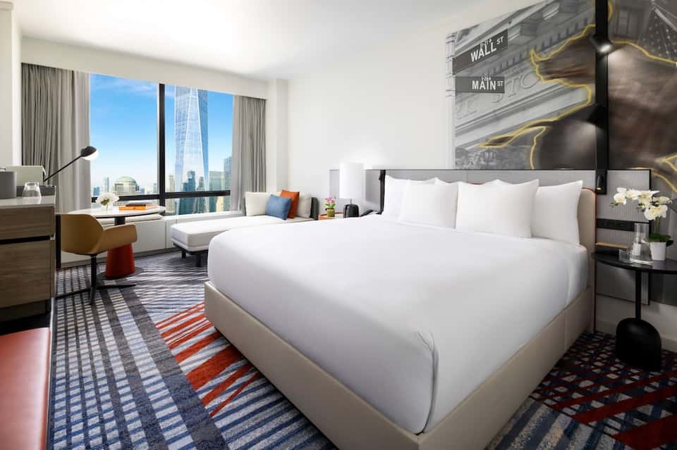High-rise hotel room with queen bed, city skyline view through floor-to-ceiling windows, and modern furnishings