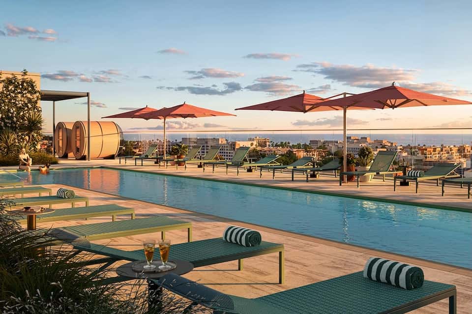 Rooftop pool deck at sunset with loungers, umbrellas, barrel sauna, and city skyline views beyond