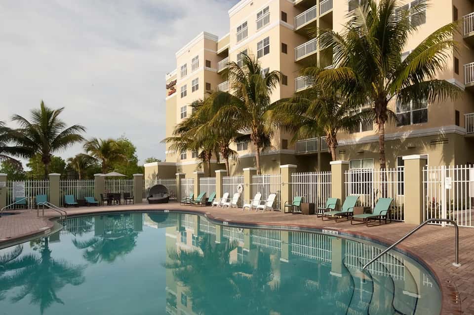 Resort pool area with loungers, white fencing, multi-story hotel building, and palm trees