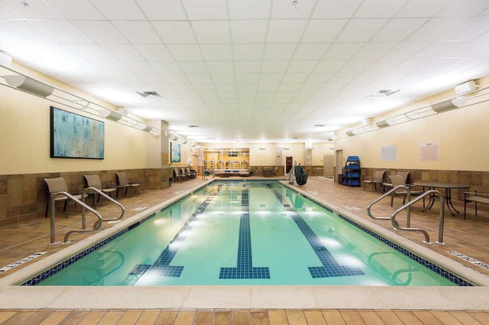 Indoor lap pool with lane markings, seating area, and bright fluorescent lighting in fitness facility