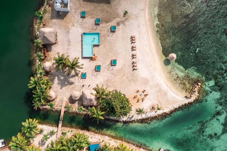 Aerial beach view with turquoise pool, lounge chairs, palm trees, and clear green ocean water