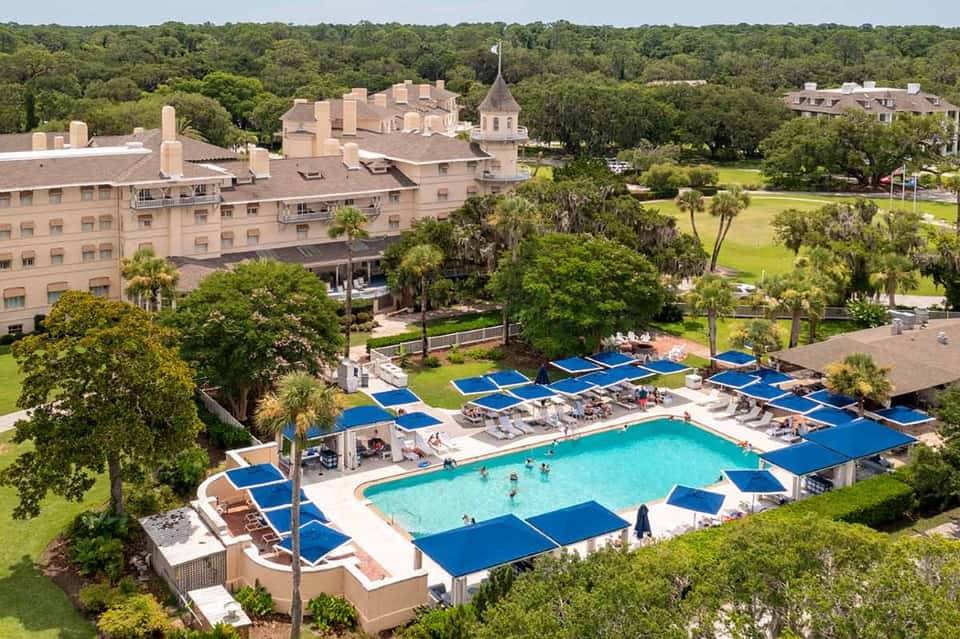Aerial view of resort pool with blue umbrellas, surrounding grounds, and historic buildings