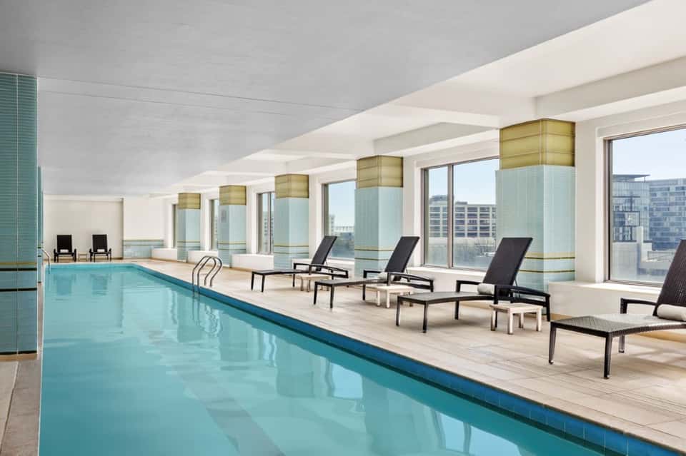 Indoor lap pool with floor-to-ceiling windows, lounge chairs, and city skyline views