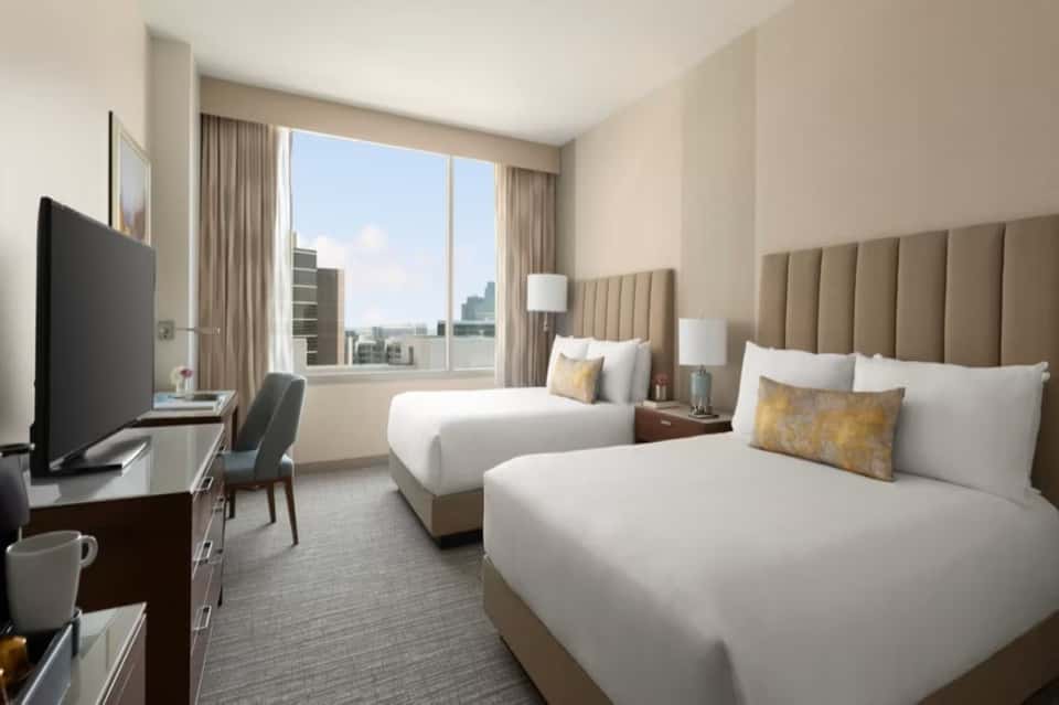 Modern hotel guest room with two queen beds, city view windows, TV, and contemporary furnishings
