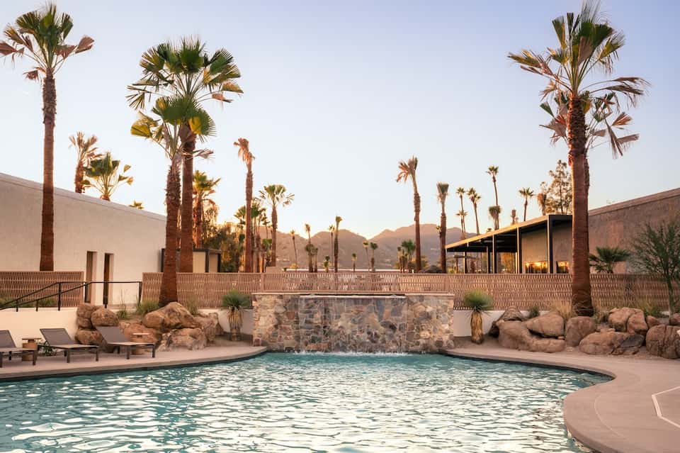 Desert resort pool surrounded by tall palms, stone waterfall feature, contemporary architecture, and mountains