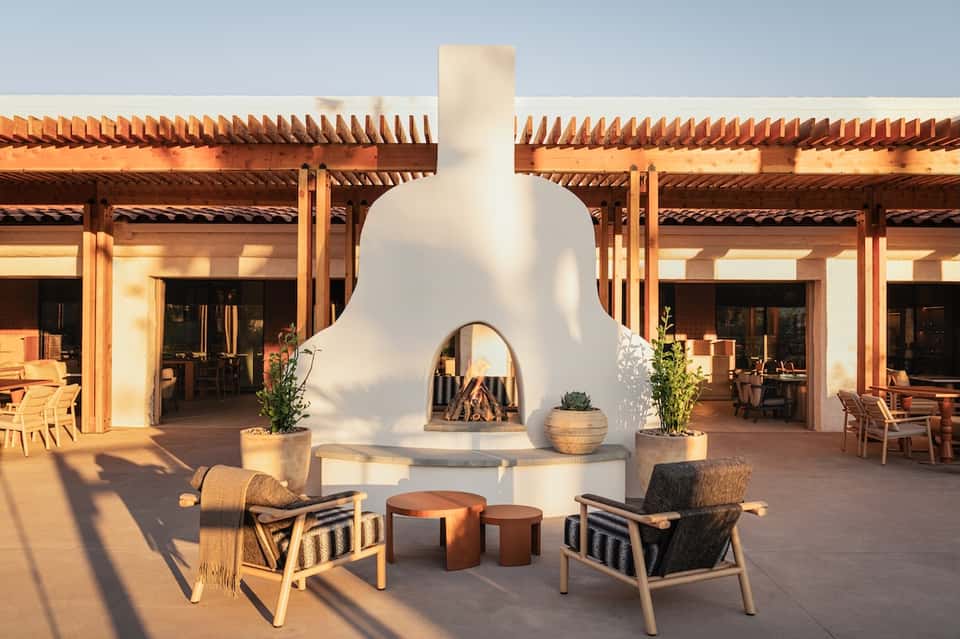 Desert resort courtyard with sculptural white fireplace, wooden pergola, natural seating, and sunset lighting