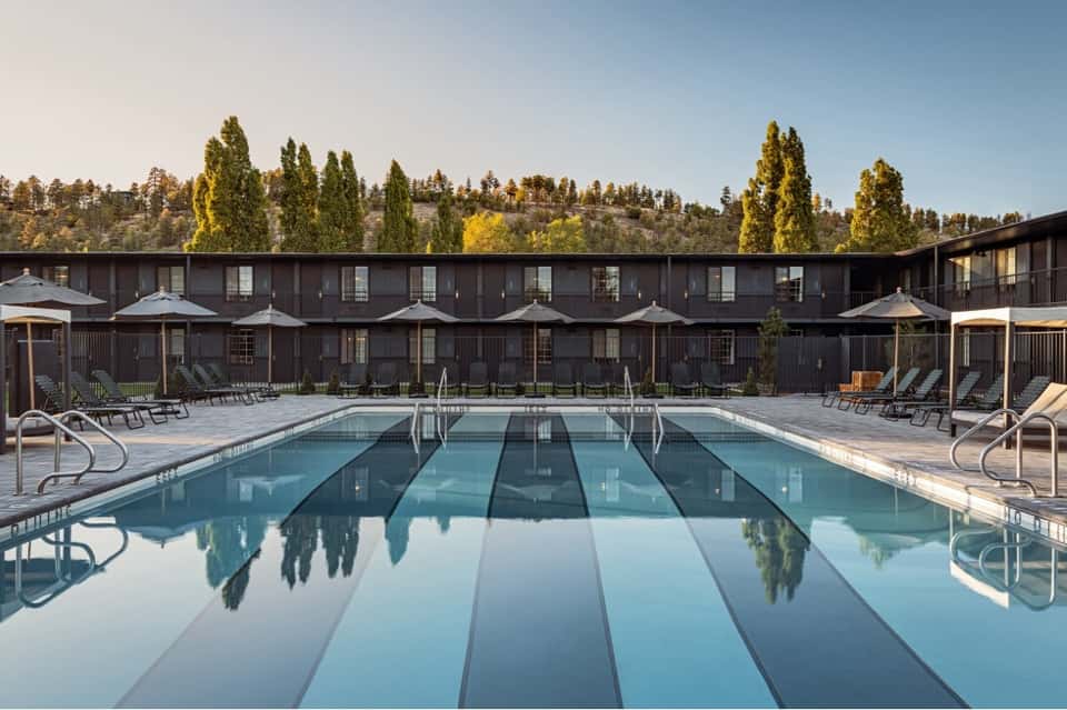 Olympic-size outdoor swimming pool with lounge chairs, modern resort building, and pine forest backdrop