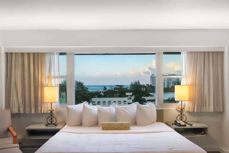 Hotel bedroom with ocean view windows, white bedding, elegant lamps, and coastal scenery