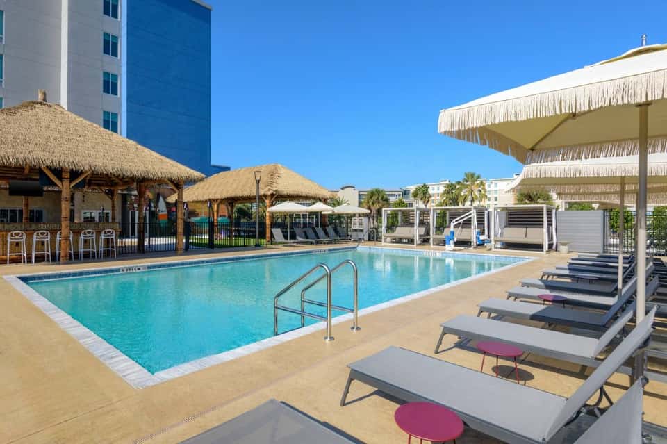 Resort pool deck with thatched-roof bar, lounge chairs, umbrellas, and modern high-rise hotel building