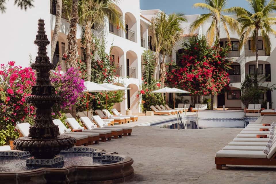 Luxury resort courtyard with white arched architecture, vibrant bougainvillea flowers, lounge chairs, and decorative fountain.