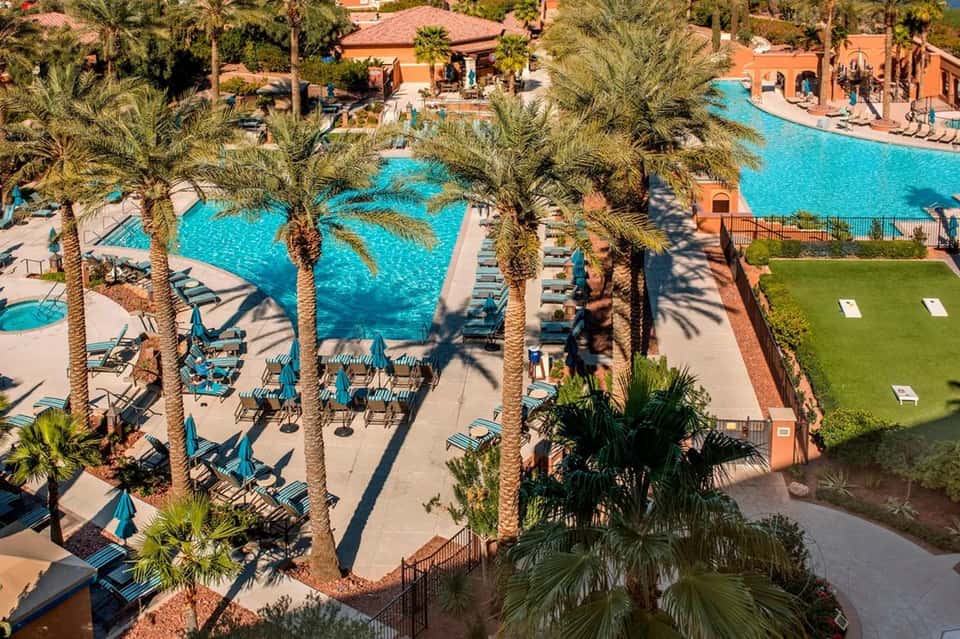 Aerial view of large resort pool complex with palm trees, lounge chairs, and terracotta buildings