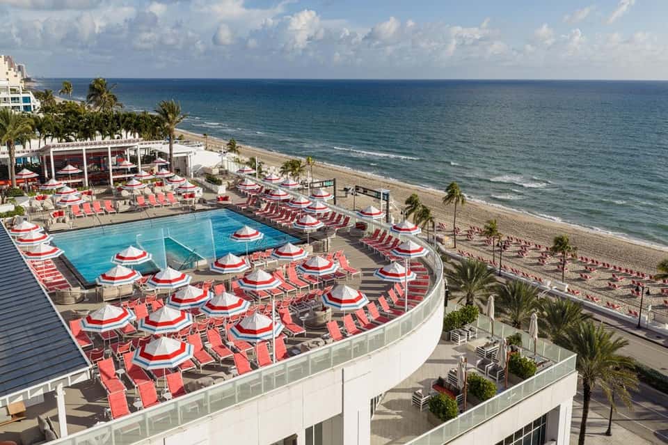 Resort pool with red and white umbrellas overlooking beach and palm trees
