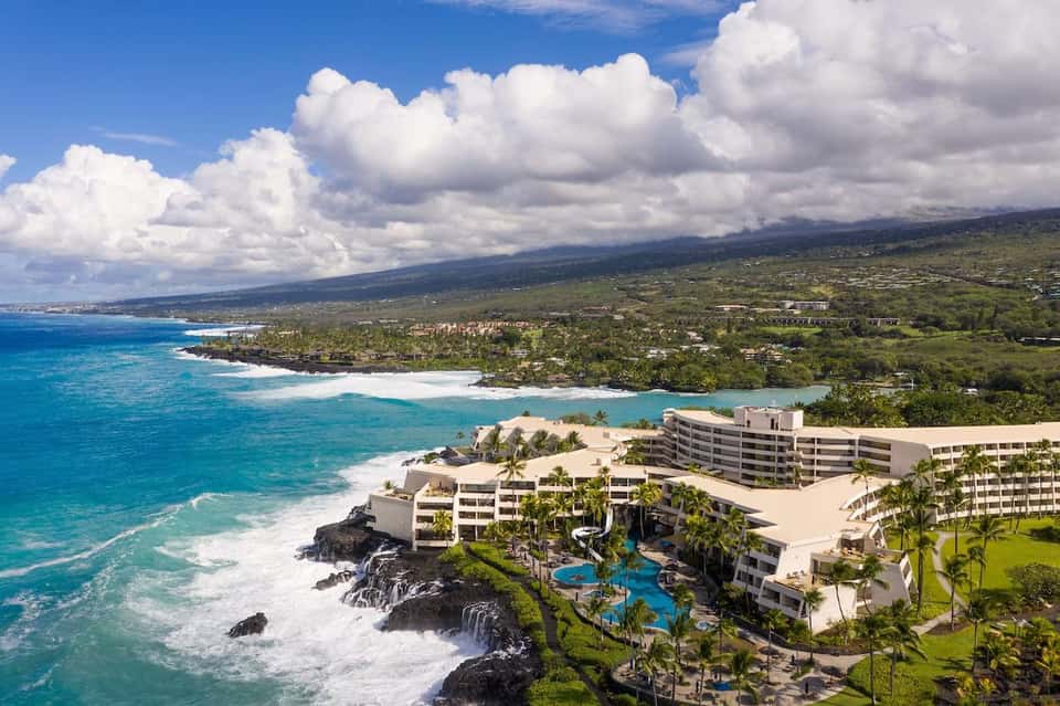 Aerial resort view featuring pools, buildings, palm trees, and dramatic coastline landscape