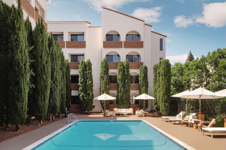 Hotel courtyard with pool, white umbrellas, lounge chairs, cypress trees, and multi-story cream building with wooden accents