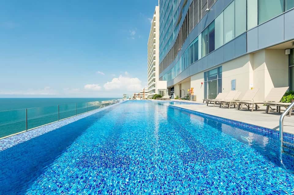 Infinity pool overlooking the turquoise ocean with high-rise hotel building and lounge chairs