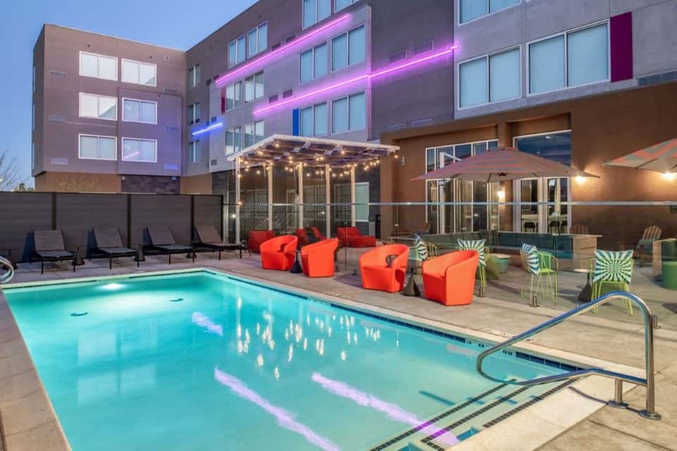 Contemporary hotel exterior with pool, red lounge chairs, pink neon lighting on building facade, and modern architecture