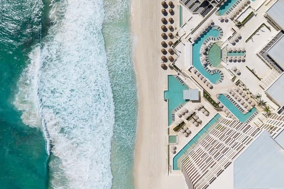 Aerial view of resort pools, beach loungers, and umbrellas along sandy coastline with ocean waves