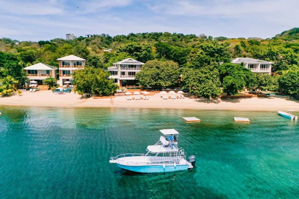 Beachfront resort villas with turquoise ocean, white boats, sandy beach, and lush hillside backdrop