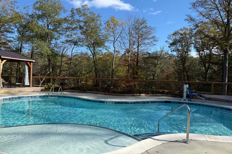 Crystal-clear resort pool with hot tub, wooden deck, gazebo, and autumn forest landscape.