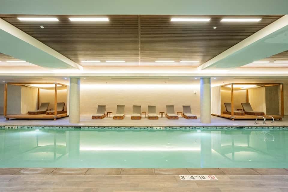 Indoor lap pool with turquoise water, lounge chairs, and relaxation alcoves with wooden canopies