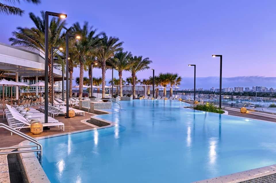 Illuminated infinity pool at twilight with palm trees, lounge chairs, and city skyline views