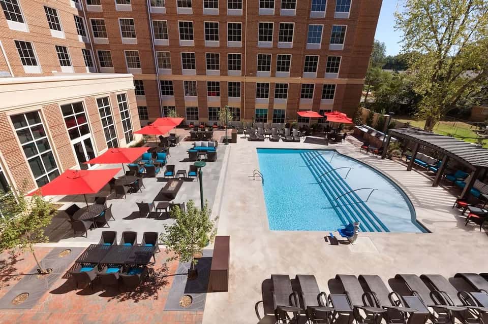 Outdoor pool area with red umbrellas, lounge chairs, and multi-story brick hotel building