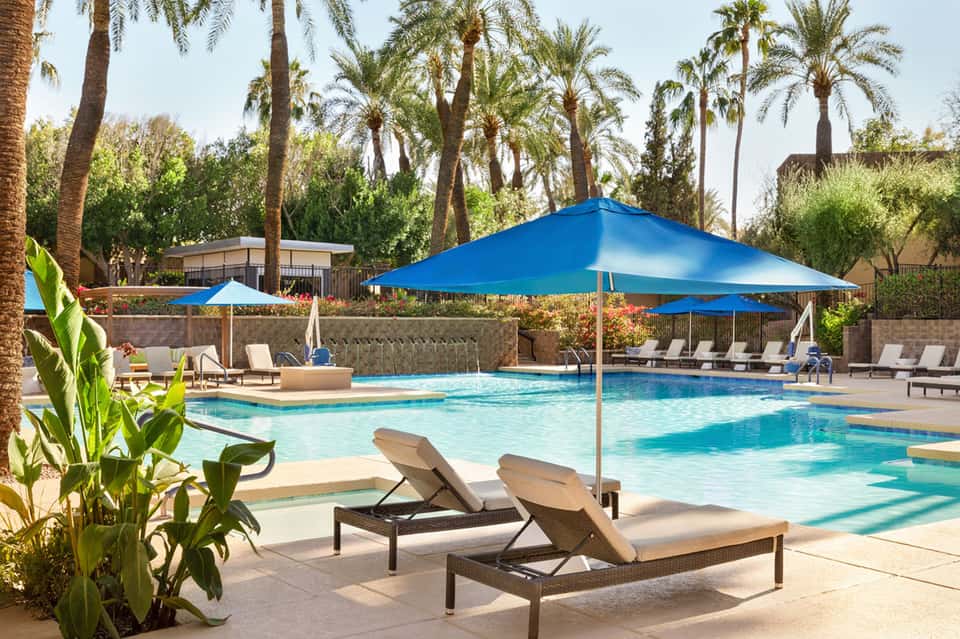 Resort pool surrounded by palm trees, blue umbrellas, lounge chairs, and lush landscaping under clear sky