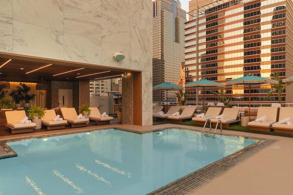 Indoor heated pool with turquoise water, lounge chairs, and city skyline views from hotel terrace