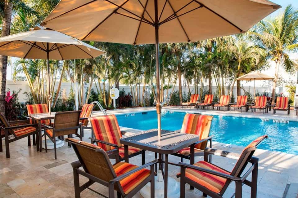Resort poolside with blue swimming pool, striped lounge chairs, tan umbrellas, and palm trees