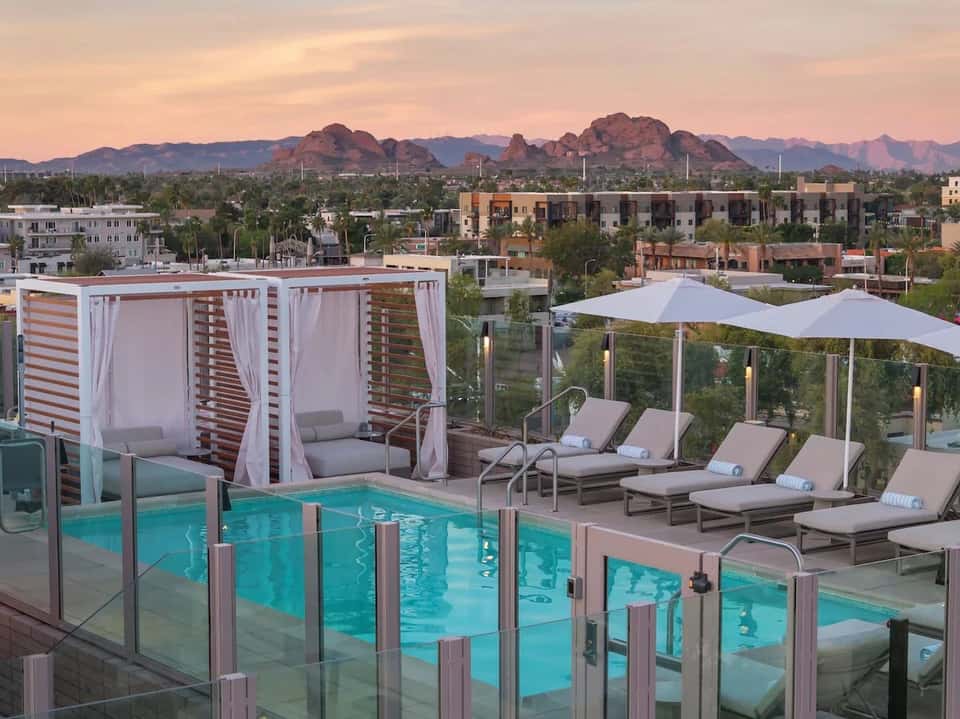 Rooftop pool at sunset with cabanas, loungers, white umbrellas, and mountain views