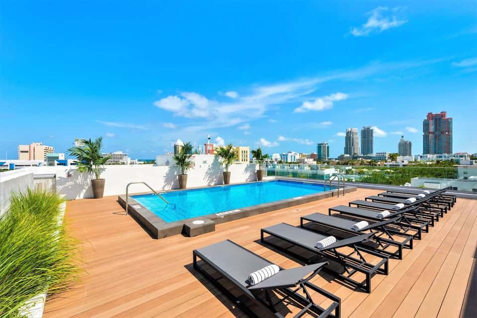 Rooftop swimming pool with lounge chairs, ocean view, palm trees, and modern city skyline in background