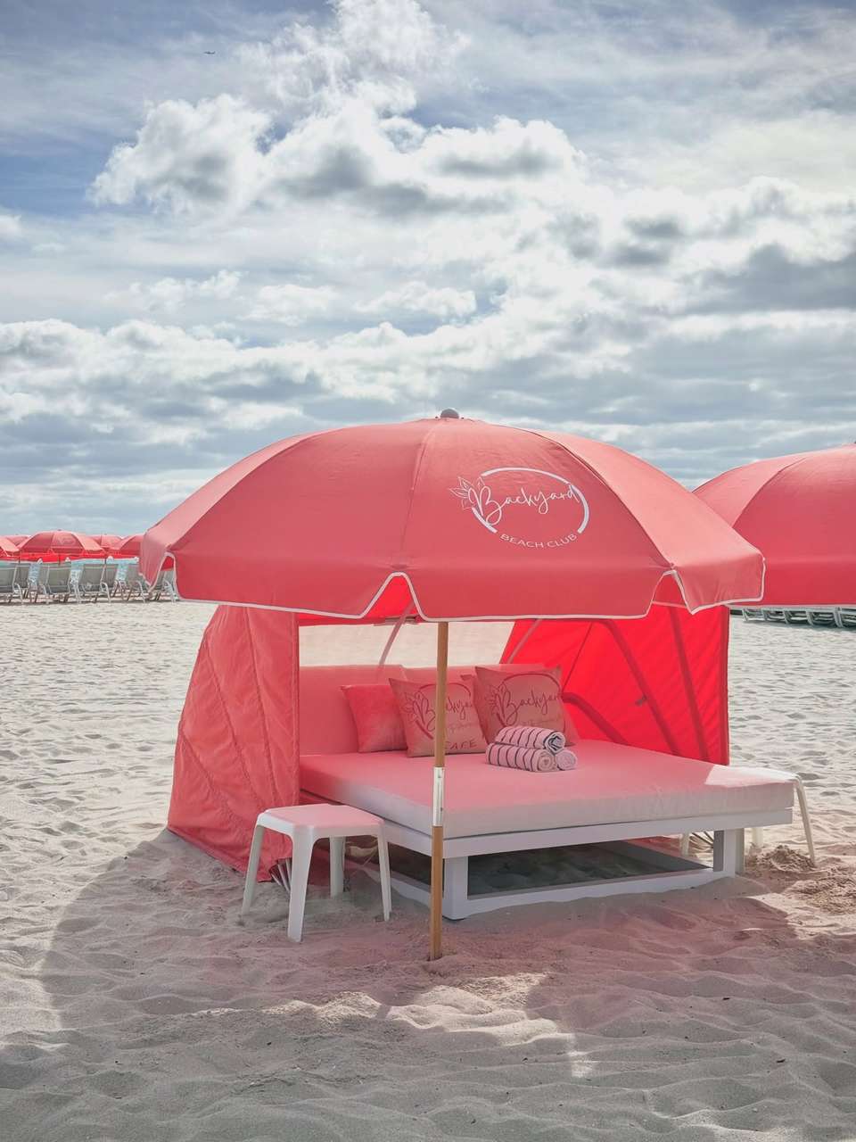 Pink cabana tent on beach with lounge bed, umbrella, pillows, and clouds visible in sky