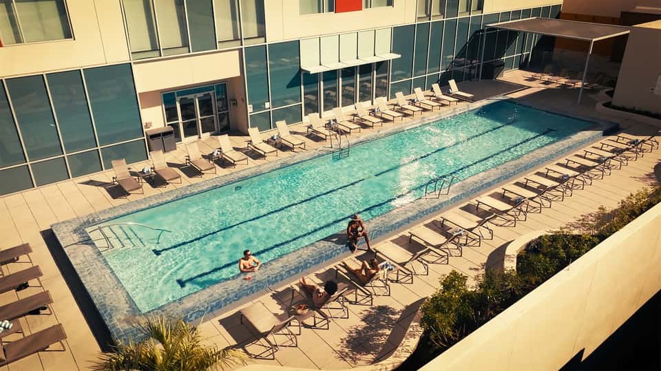Indoor lap pool with lane markers and lounge chairs along glass-walled facility in bright daylight