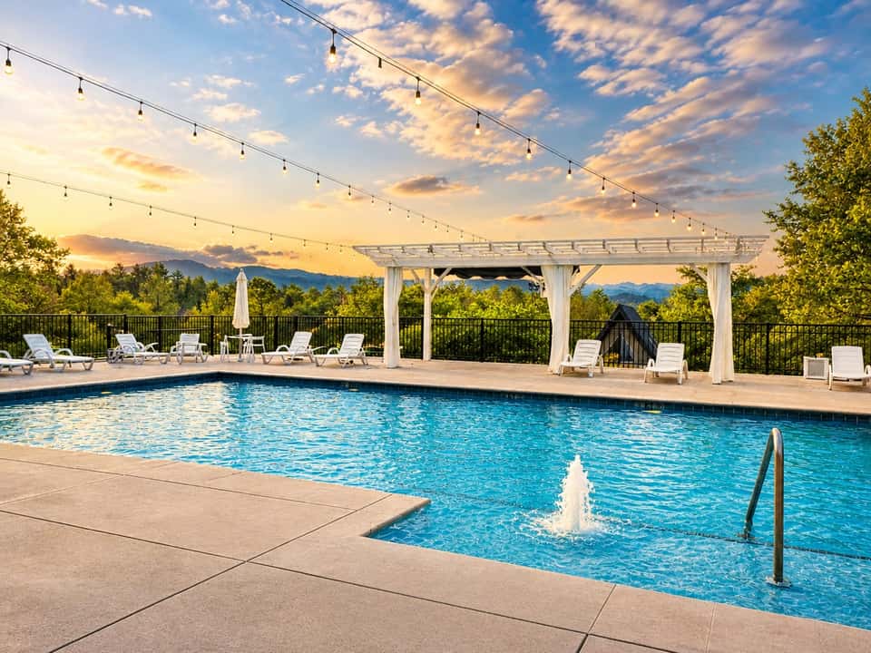 Resort pool with pergola, string lights, lounge chairs, and mountain views at sunset