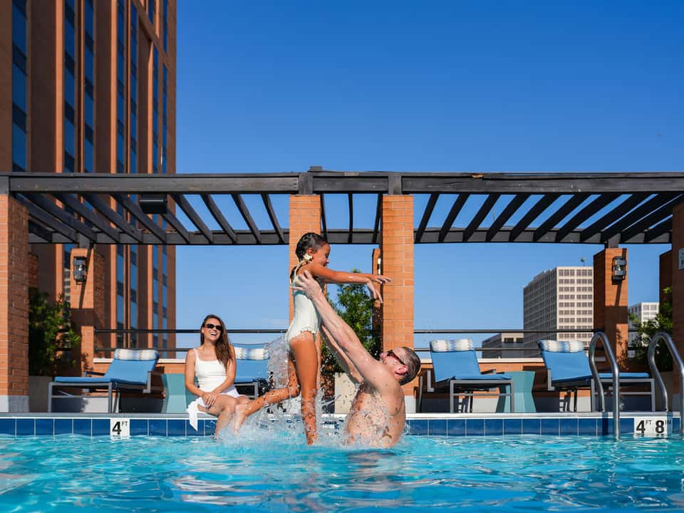 Family playing in hotel pool with lounge chairs on deck and city buildings in background